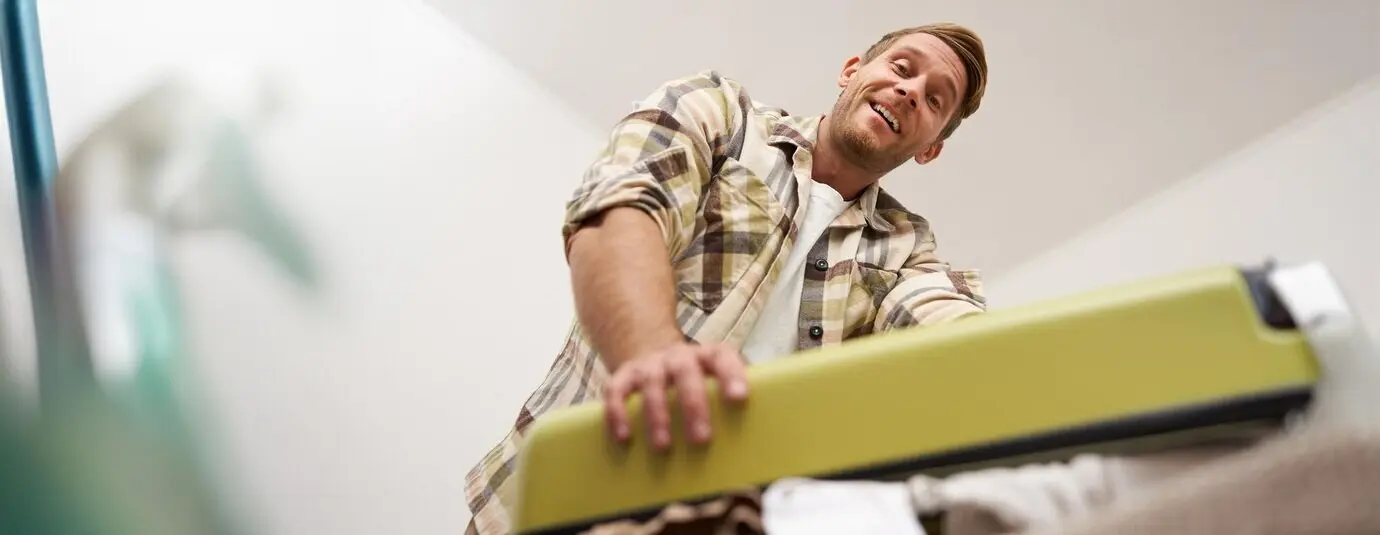 Low-angle portrait of a muscular young man trying to close his suitcase full of clothes, pressing down on the bag with effort.